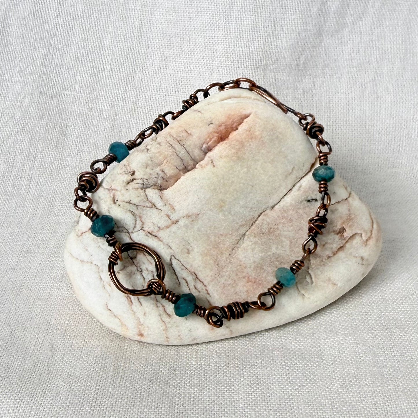 A handmade copper wire-wrapped bracelet with small teal beads, displayed on a polished stone against a textured off-white fabric background.