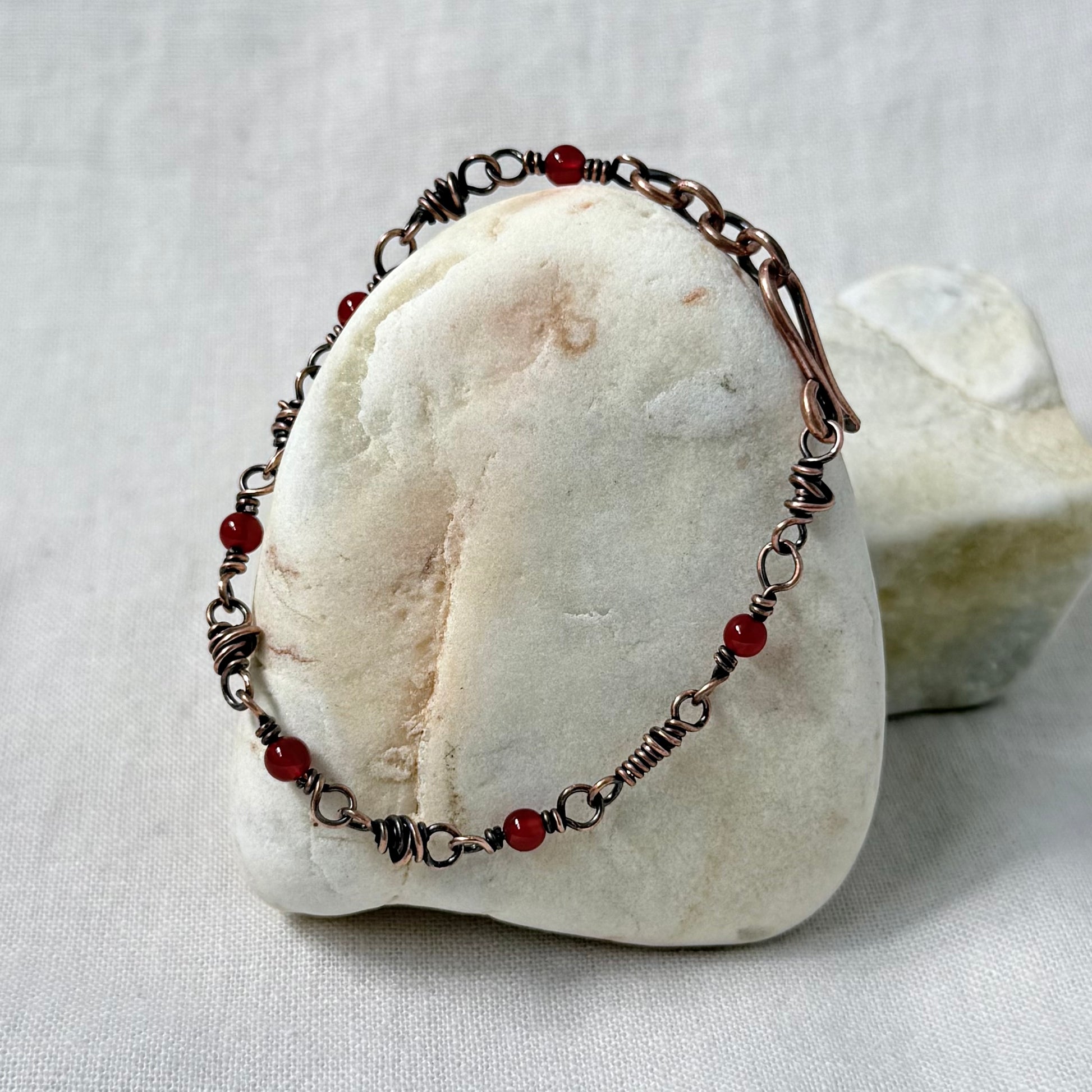 A handmade copper wire-wrapped bracelet with small red beads, displayed on a white stone stand against a light fabric background.