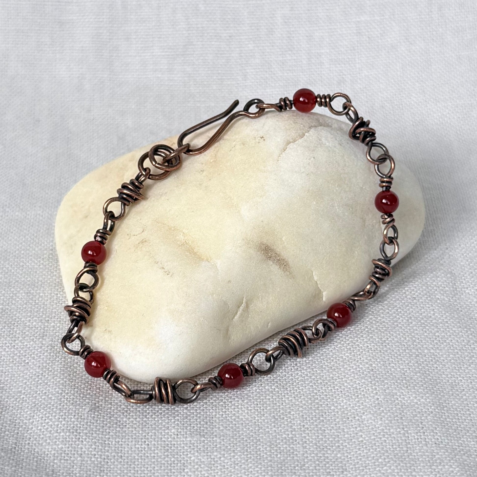 A handmade copper wire-wrapped bracelet with small red carnelian beads, displayed on a smooth white stone against a textured white fabric background.