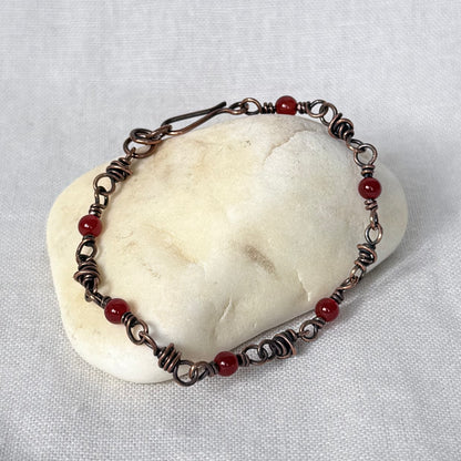 A handmade wire-wrapped bracelet with small red carnelian beads, displayed on a smooth white stone against a textured white fabric background.
