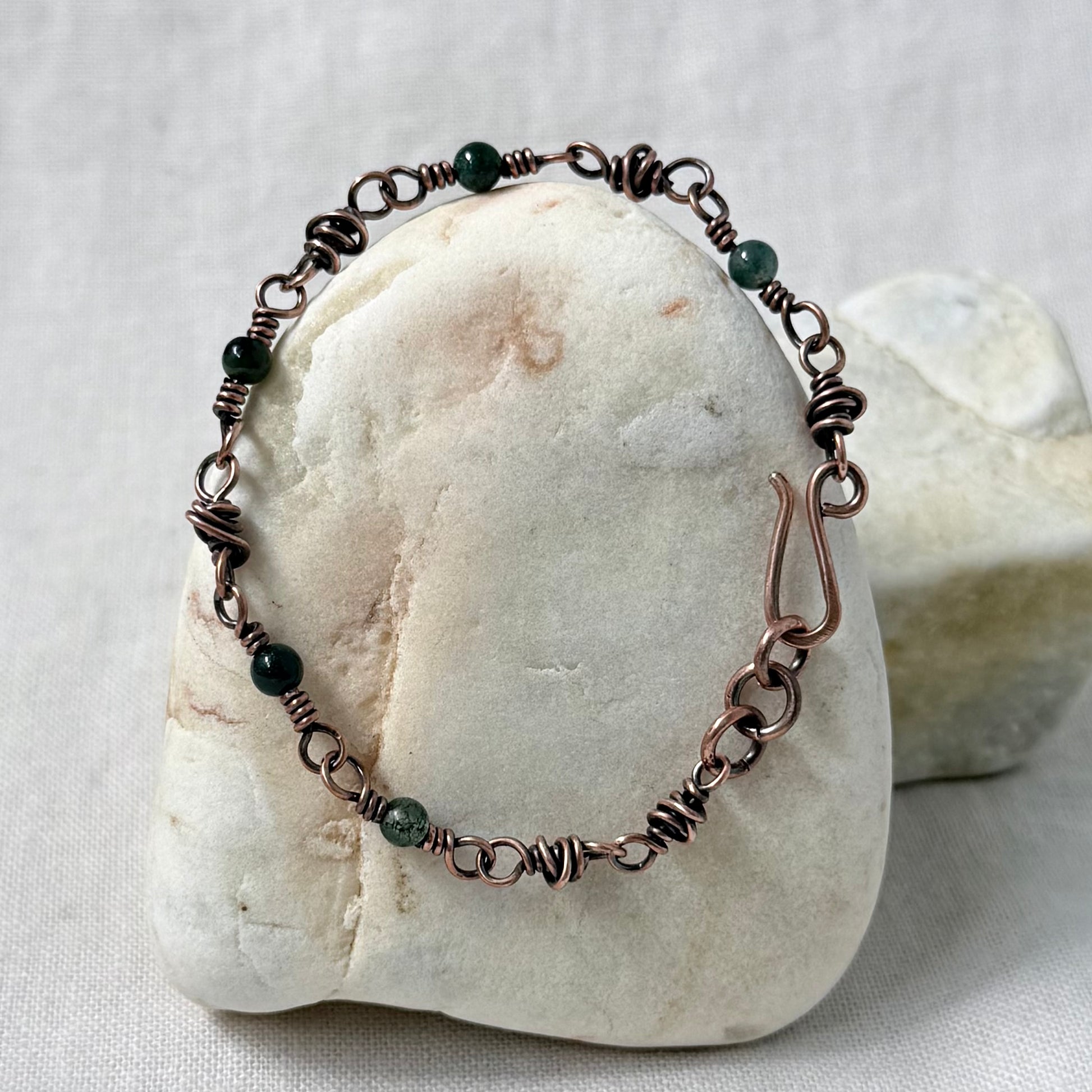 A handmade copper wire wrapped bracelet with dark green beads, displayed on a smooth white stone against a neutral fabric background.
