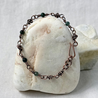 A handmade copper wire wrapped bracelet with dark green beads, displayed on a smooth white stone against a neutral fabric background.