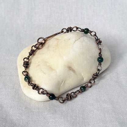 A handmade copper wire-wrapped bracelet with dark green Moss Agate beads, displayed on a smooth, light-coloured stone against a textured white fabric background.