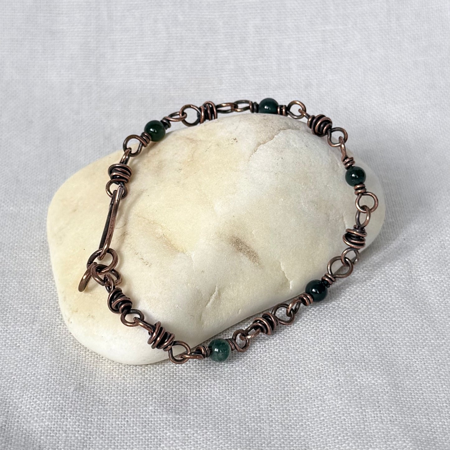 A handmade copper wire bracelet with dark green Moss Agate beads, displayed on a smooth, rounded beige stone against a textured light grey fabric background.