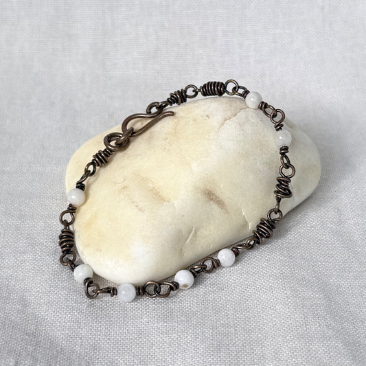 A handmade copper bracelet with white round moonstone beads and copper wire wrapping, displayed on a smooth cream-colored stone against a textured light gray fabric background.