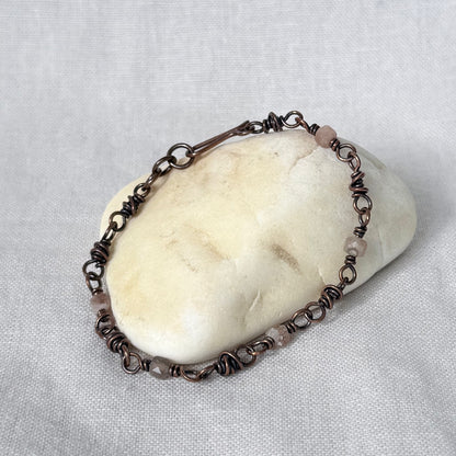 A handmade copper link bracelet with small dusty sunstone beads, displayed on a smooth cream-coloured stone against a textured light grey fabric background.