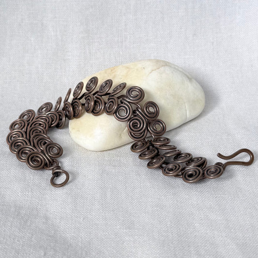 A copper chain bracelet with intricate spiral curls and small hooks, draped over a smooth white stone on a textured light grey fabric background.
