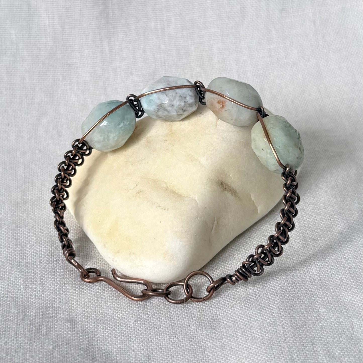 A handmade copper wire bracelet with four light blue-green stone beads, displayed on a cream-coloured stone stand against a textured white fabric background.