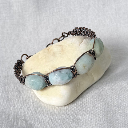 A handmade copper wire-wrapped bracelet featuring light blue-green amazonite stones, displayed on a small white stone against a neutral grey fabric background.