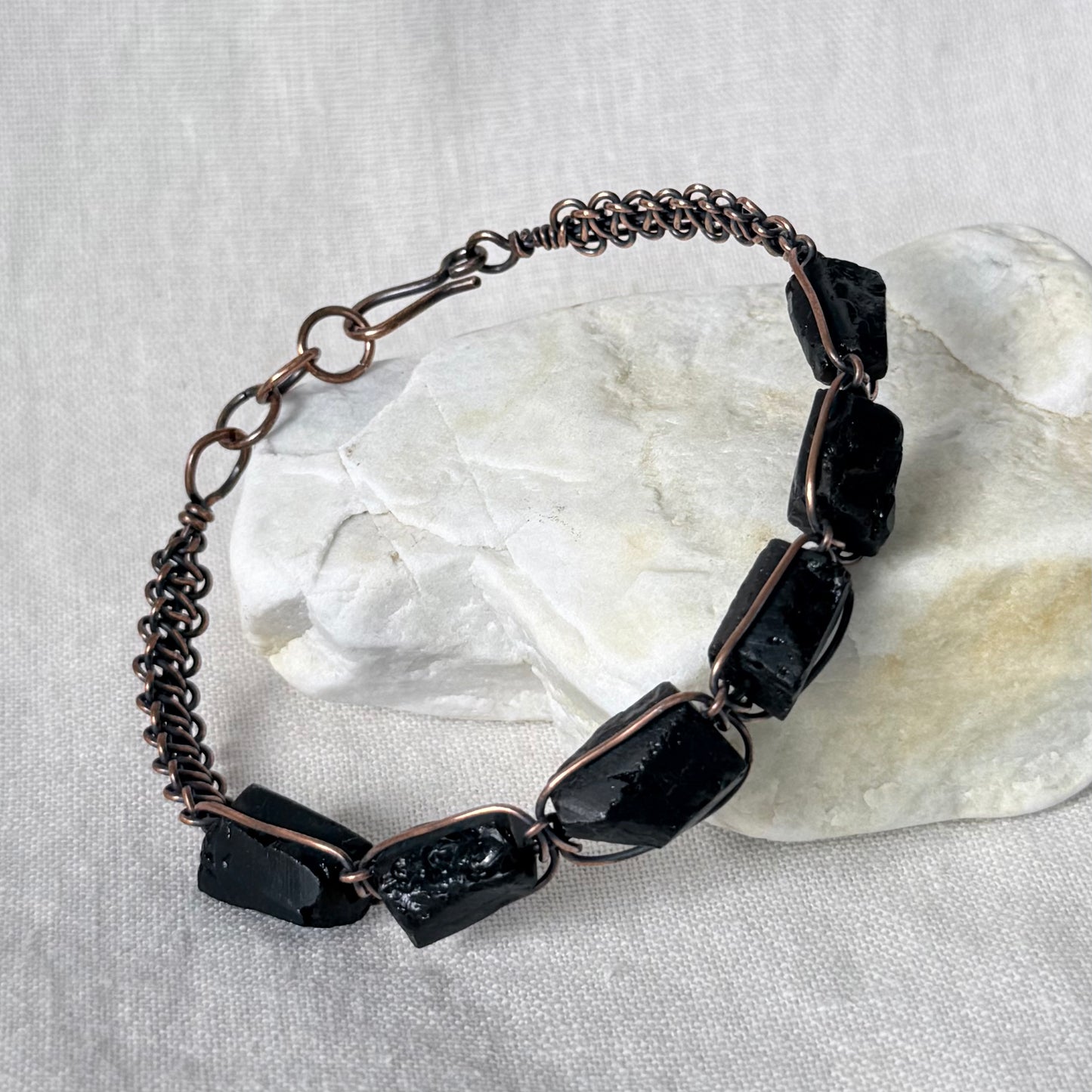 A handmade bracelet featuring raw black stones set in copper, displayed on a white textured fabric background with a white decorative stone prop.