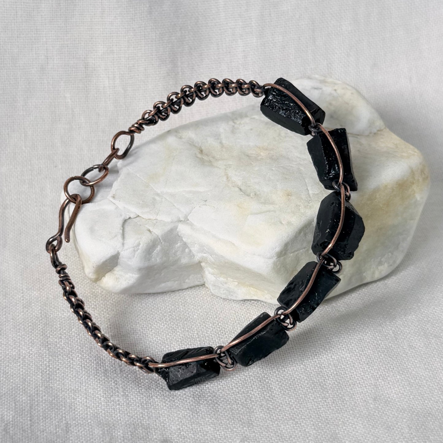 A handmade copper bracelet featuring dark, polished rectangular stone beads, displayed on a white textured fabric background with a white stone prop.