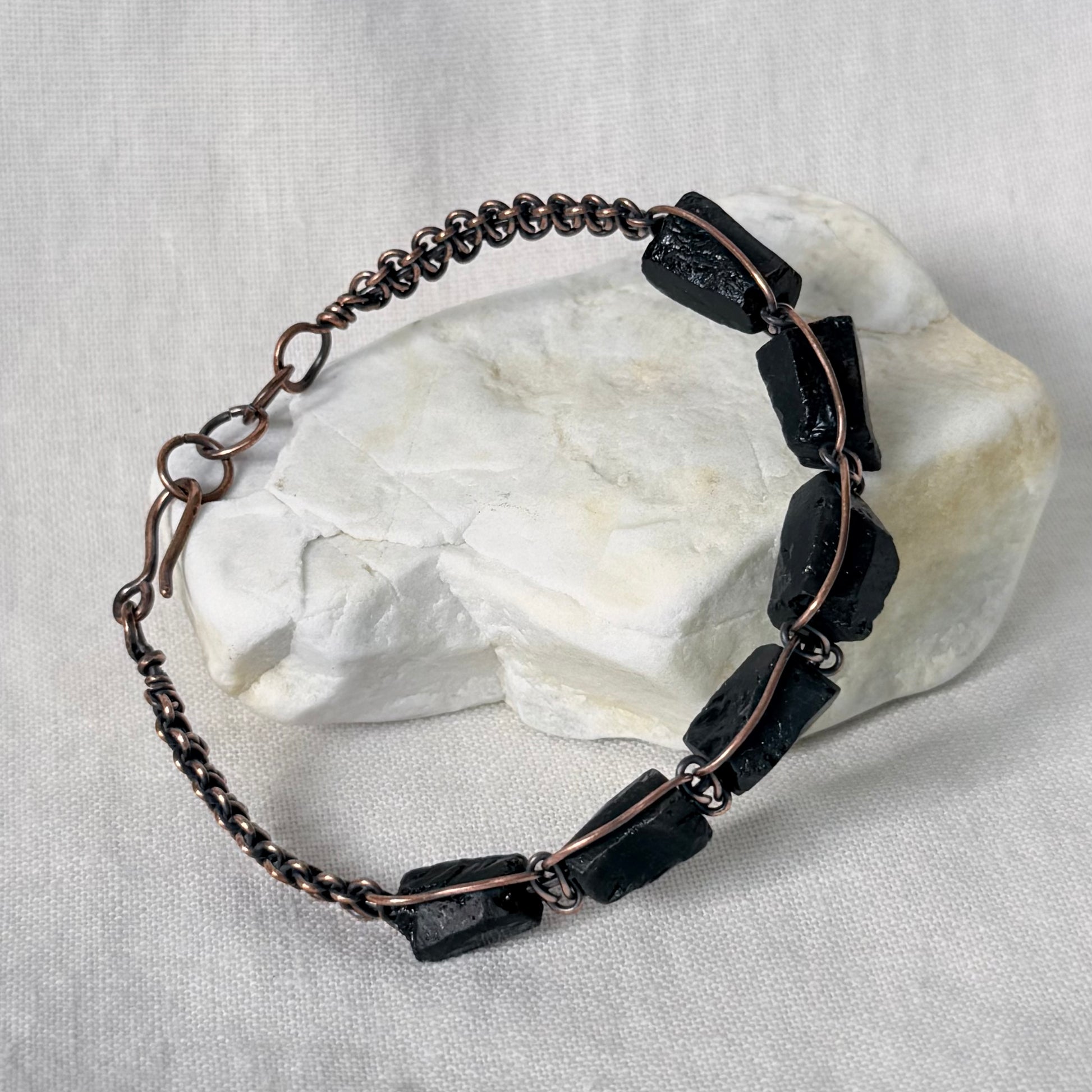 A handmade copper bracelet featuring dark, polished rectangular stone beads, displayed on a white textured fabric background with a white stone prop.