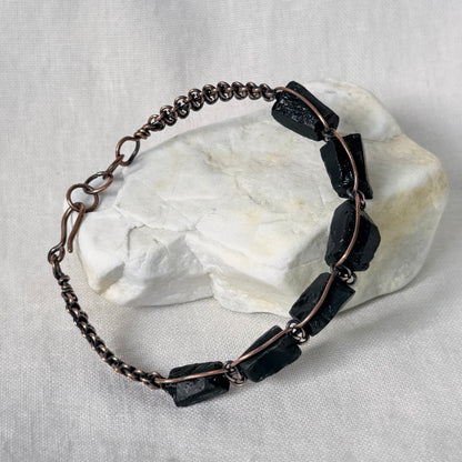 A handmade copper bracelet featuring dark, polished rectangular stone beads, displayed on a white textured fabric background with a white stone prop.