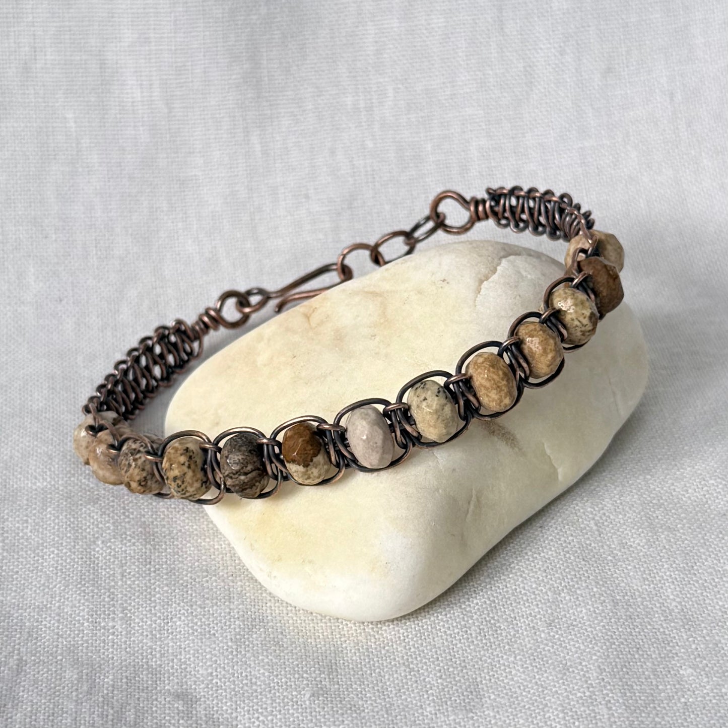 A handmade copper wire-wrapped bracelet with natural stone beads, displayed on a smooth white stone against a textured light grey fabric background.
