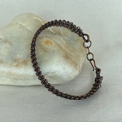 A copper woven bracelet with a coiled spring design and a spring-ring clasp, resting on a smooth white stone over a light grey fabric background.