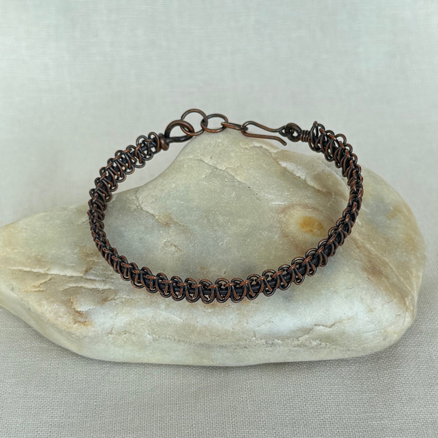A dark copper bracelet with a textured, coiled chain design and a spring-ring clasp, displayed on a smooth, light-coloured stone against a neutral fabric background.