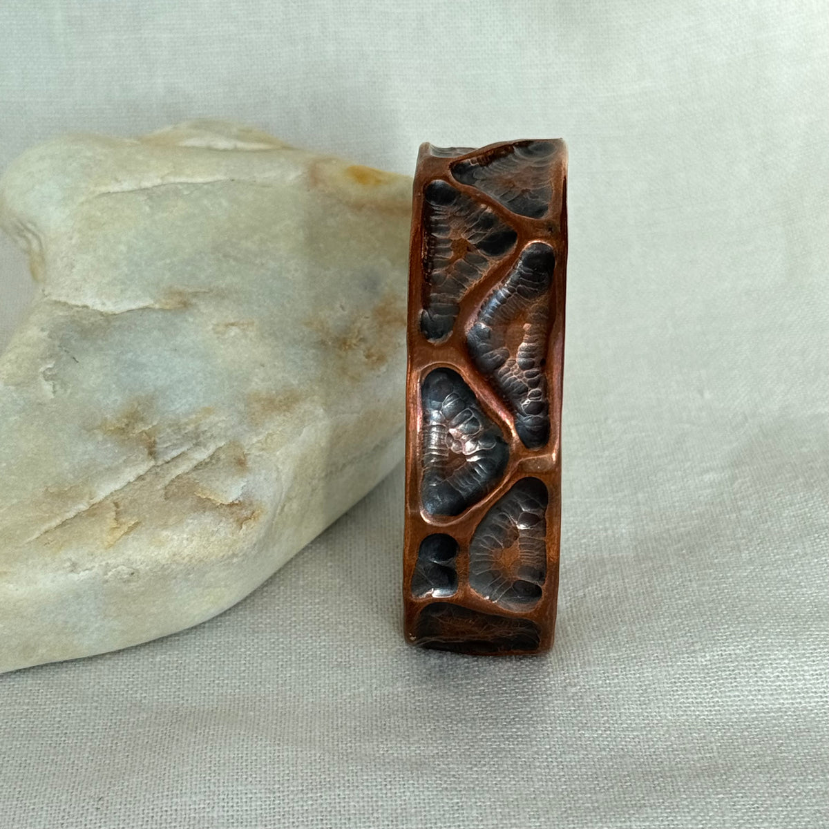 A hammered copper cuff , resting upright next to a light-coloured stone on a neutral fabric background.