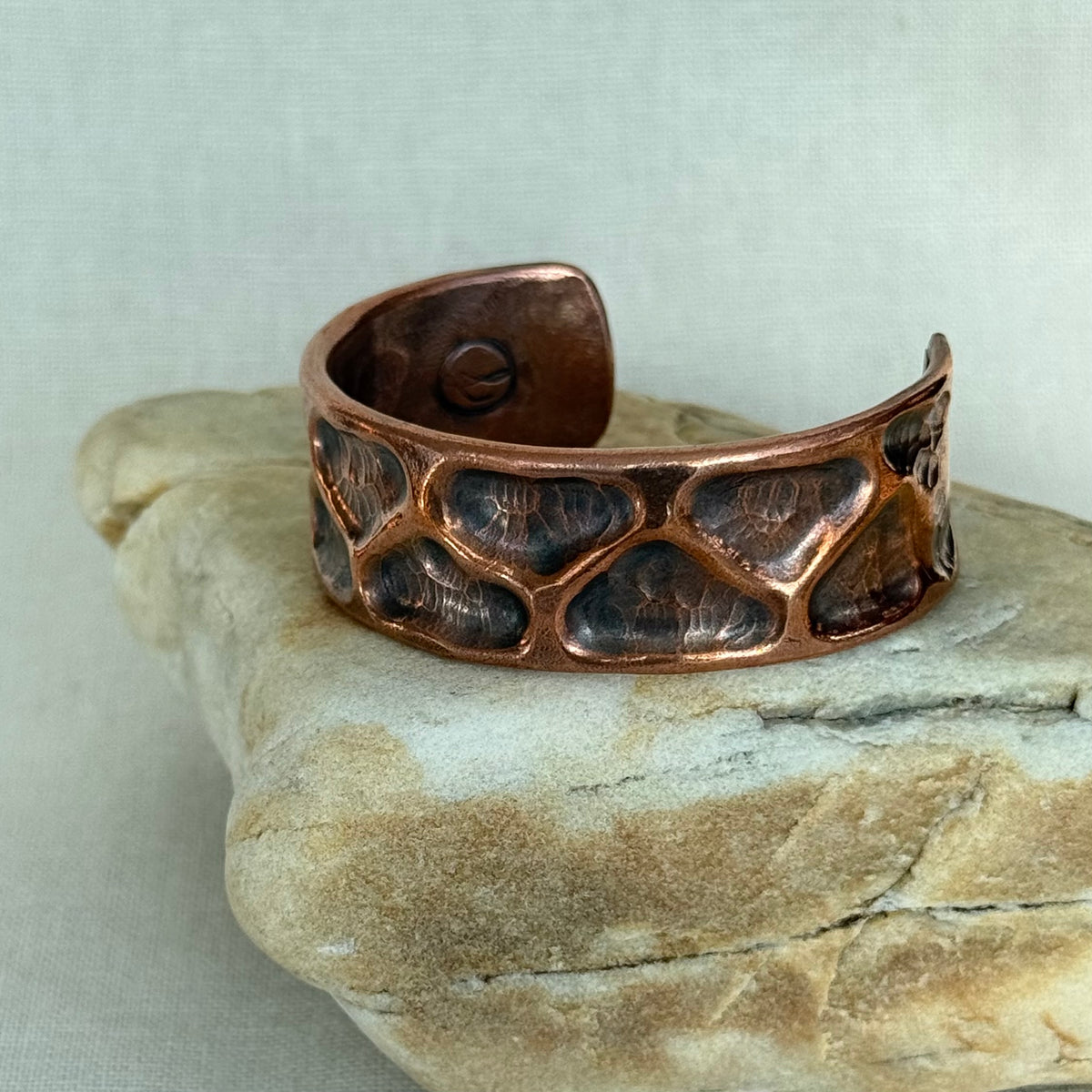 A hammered copper cuff bracelet with a textured, scale-like pattern, resting on a light-colored stone against a neutral fabric background.