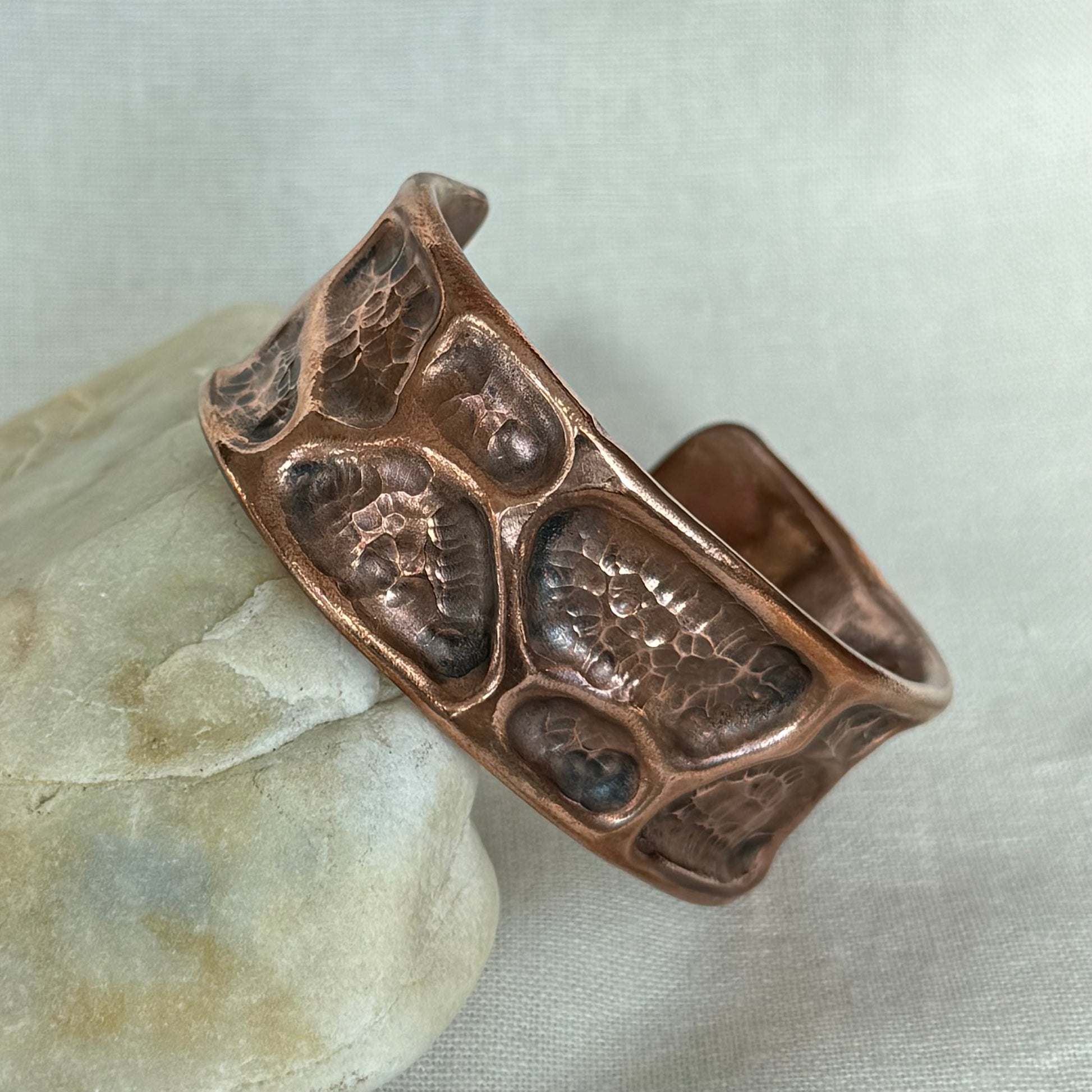 A hammered copper cuff bracelet with a textured, organic raised pattern, resting on a smooth light-colored stone against a neutral fabric background.