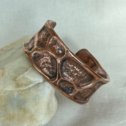 A hammered copper cuff bracelet with a textured, organic raised pattern, resting on a smooth light-colored stone against a neutral fabric background.