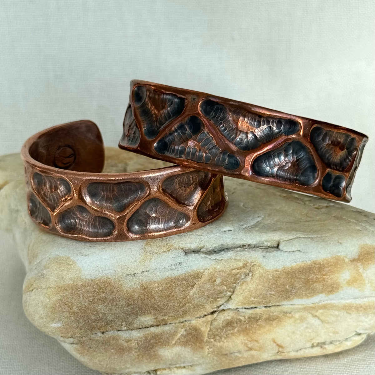 Two hammered copper cuff bracelets with a dark oxidized textured pattern, displayed on a natural stone against a neutral beige background.