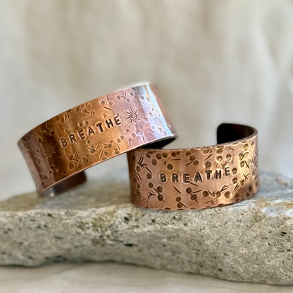 Two copper bracelets with 'BREATHE' engraving on a stone surface.