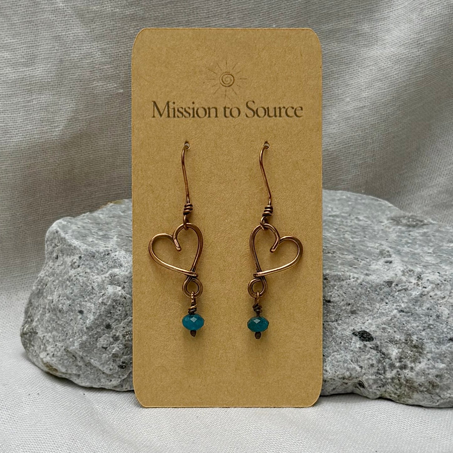 Heart-shaped copper wire earrings with bead on a 'Mission to Source' card with a stone  and linen background