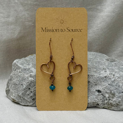 Heart-shaped copper wire earrings with bead on a 'Mission to Source' card with a stone  and linen background