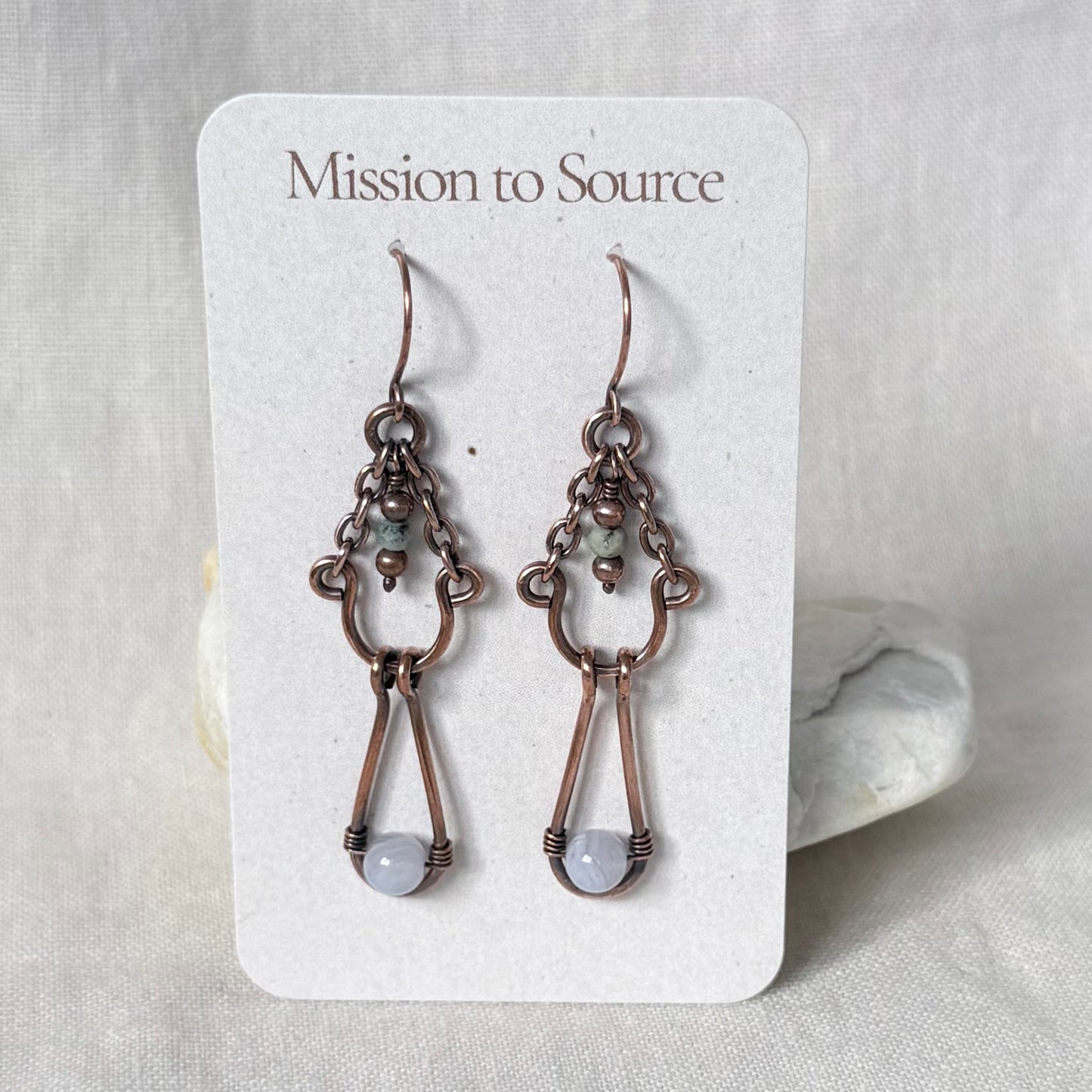 A pair of  copper handmade dangle earrings with light grey bead drops, displayed on a white card labeled 'Mission to Source' against a neutral fabric background.