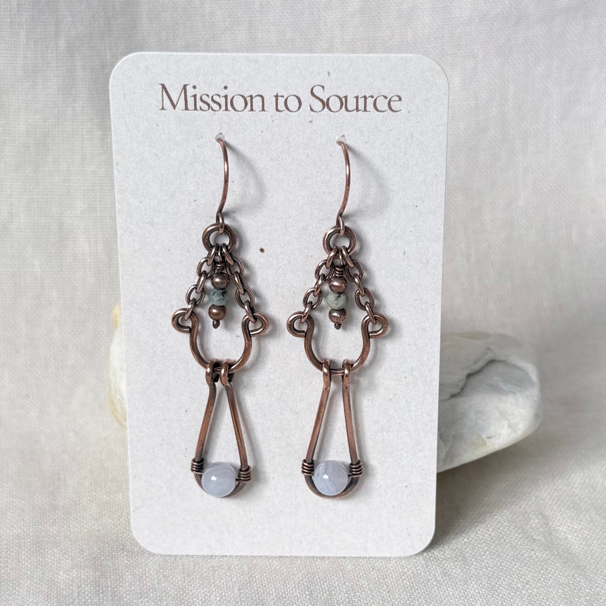 A pair of  copper handmade dangle earrings with light grey bead drops, displayed on a white card labeled 'Mission to Source' against a neutral fabric background.