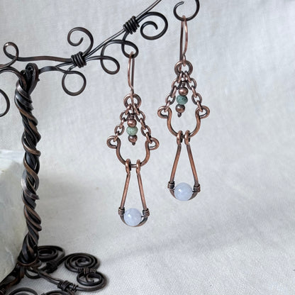 A pair of handmade copper wire drop earrings with small green beads and light blue round stones, hanging from a dark metal display stand against a white fabric background.