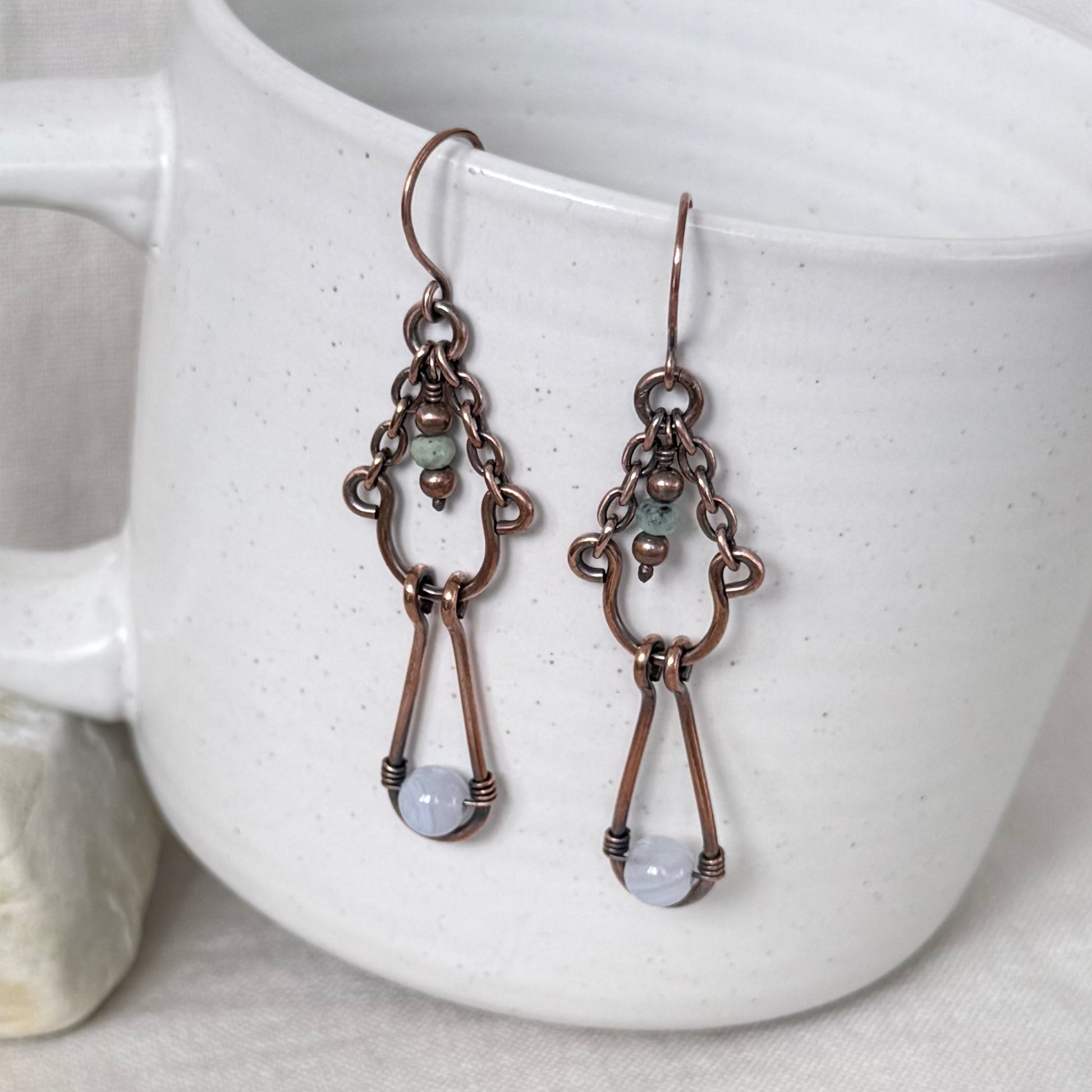 A pair of handmade copper drop earrings with small green beads and light blue-gray round stones, hanging from a white speckled ceramic mug.