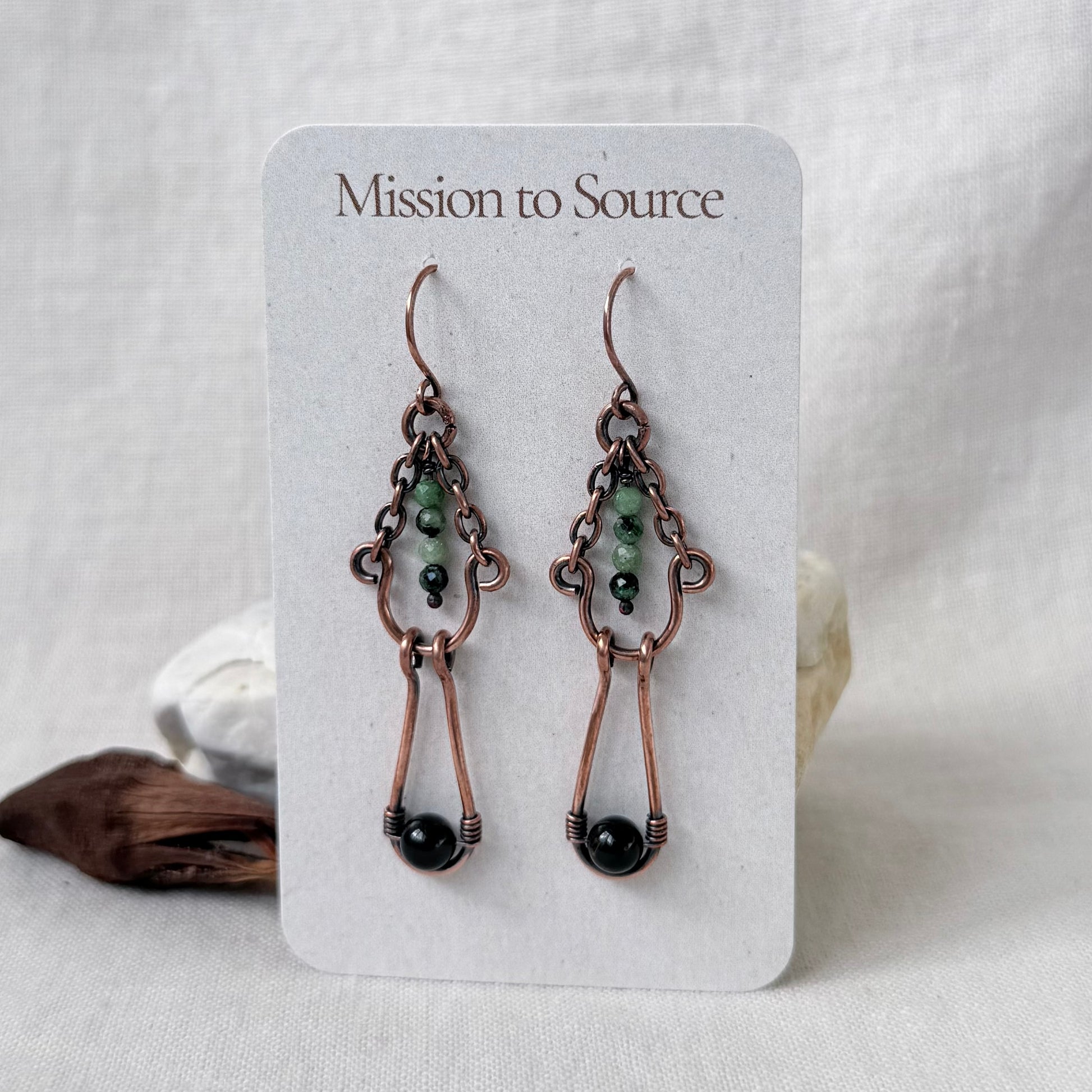 A pair of handmade copper drop earrings with green and black beads, displayed on a white card labeled 'Mission to Source'.