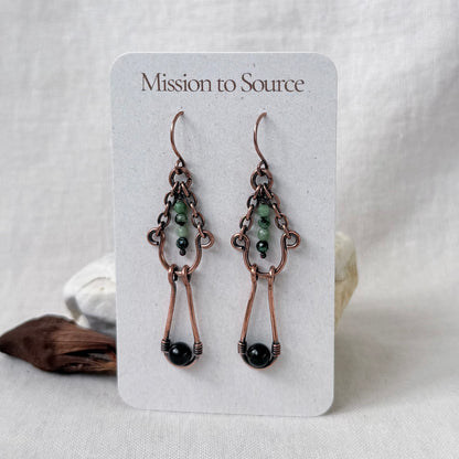 A pair of handmade copper drop earrings with green and black beads, displayed on a white card labeled 'Mission to Source'.