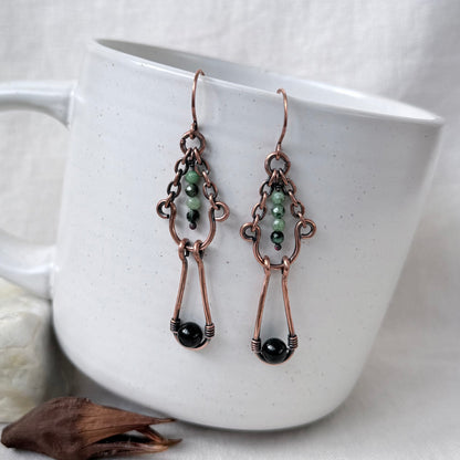 A pair of handmade copper wire drop earrings with green and black beads, hanging from a white ceramic mug.