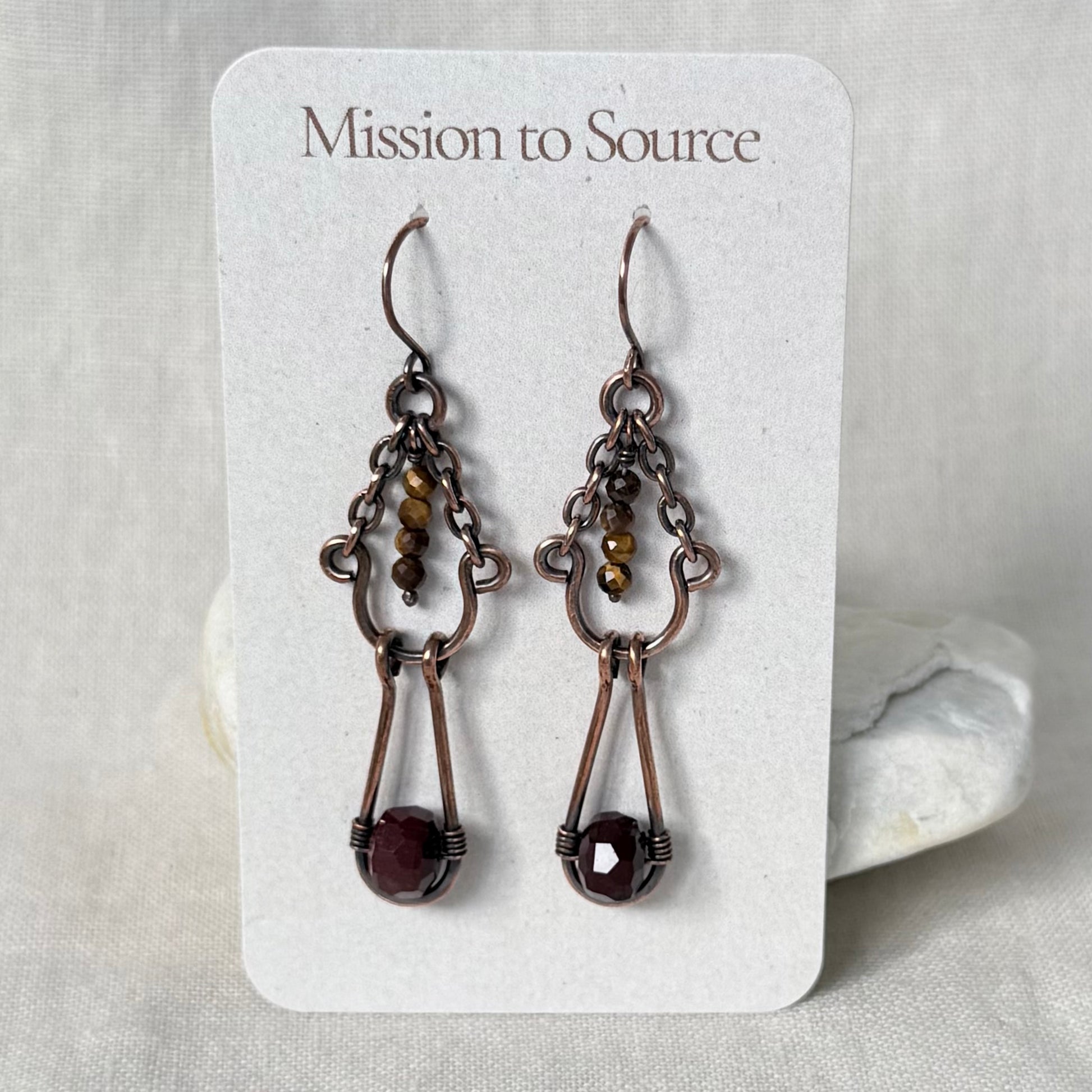 A pair of handmade copper drop earrings with dark bronze metalwork and faceted dark red gemstones, displayed on a white card labeled 'Mission to Source'.