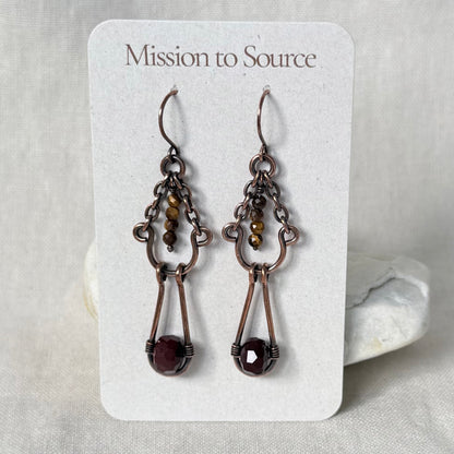 A pair of handmade copper drop earrings with dark bronze metalwork and faceted dark red gemstones, displayed on a white card labeled 'Mission to Source'.