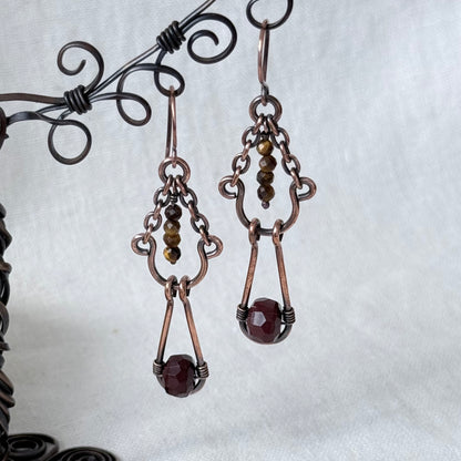 A pair of handmade copper wire drop earrings with small brown beads and faceted dark red gemstone pendants, displayed on a black wire stand against a white fabric background.