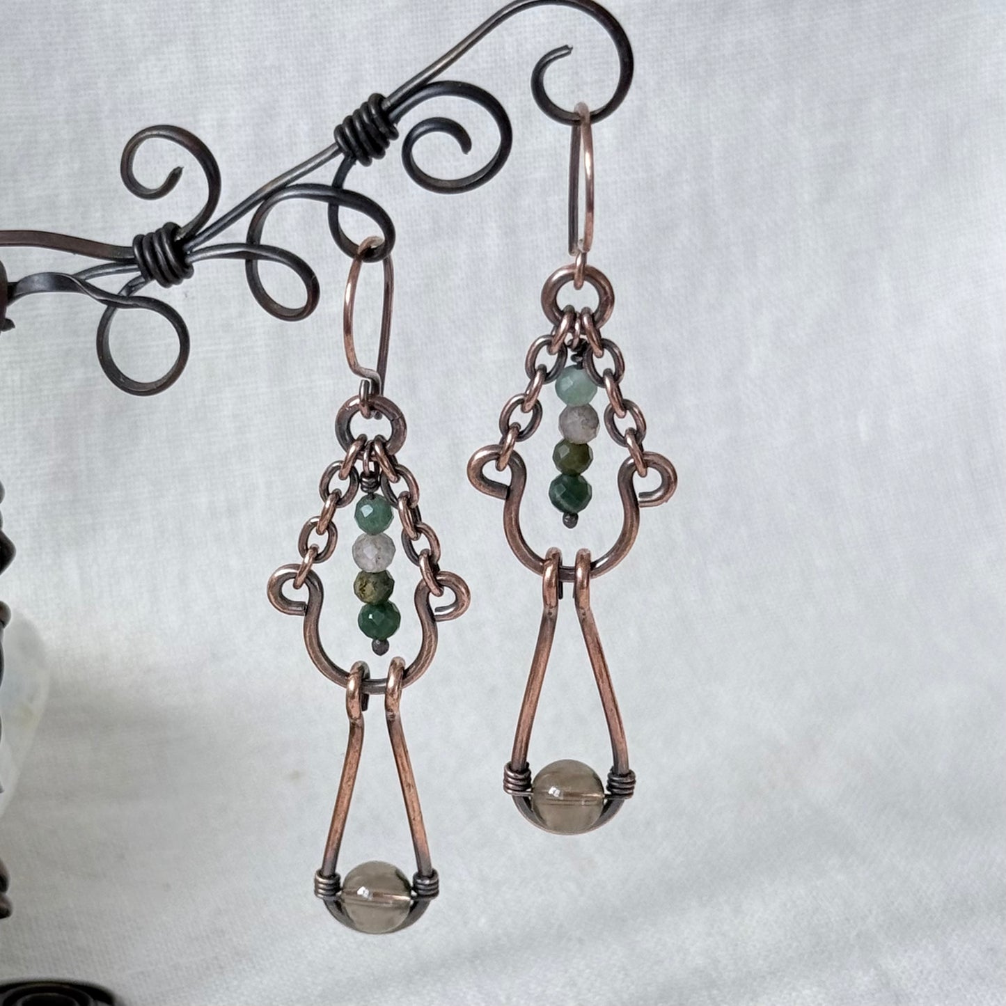 A pair of handmade copper drop earrings with green and white bead accents, hanging from a decorative black wire stand against a light gray fabric background.