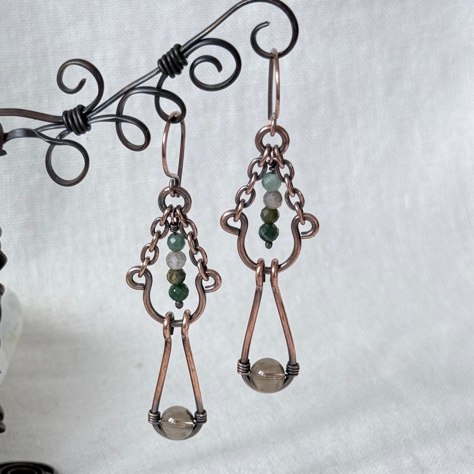 A pair of handmade copper drop earrings with green and white bead accents, hanging from a decorative black wire stand against a light gray fabric background.