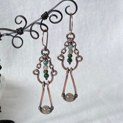 A pair of handmade copper drop earrings with green and white bead accents, hanging from a decorative black wire stand against a light gray fabric background.