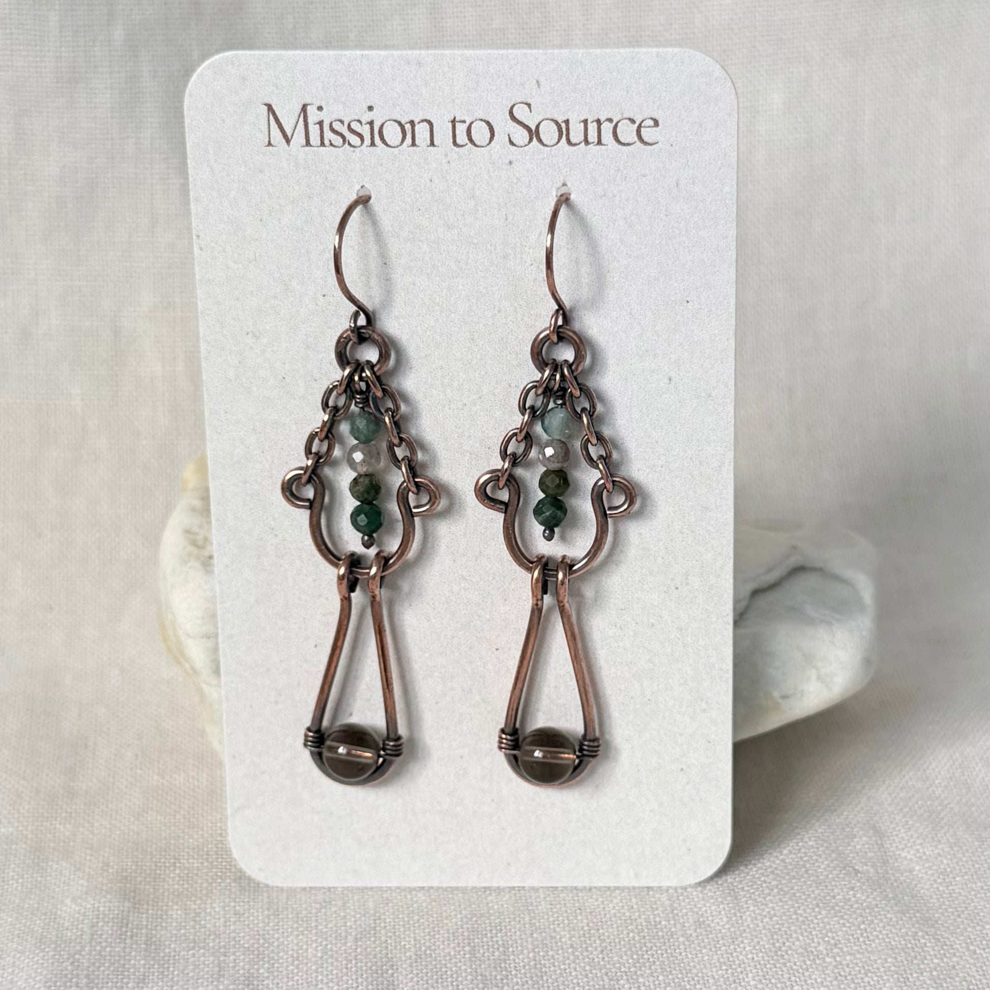 A pair of Mission to Source copper dangle earrings displayed on a white branded card. The earrings feature copper wirework with small green and white beads, set against a soft neutral fabric background.