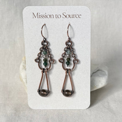 A pair of Mission to Source copper dangle earrings displayed on a white branded card. The earrings feature copper wirework with small green and white beads, set against a soft neutral fabric background.
