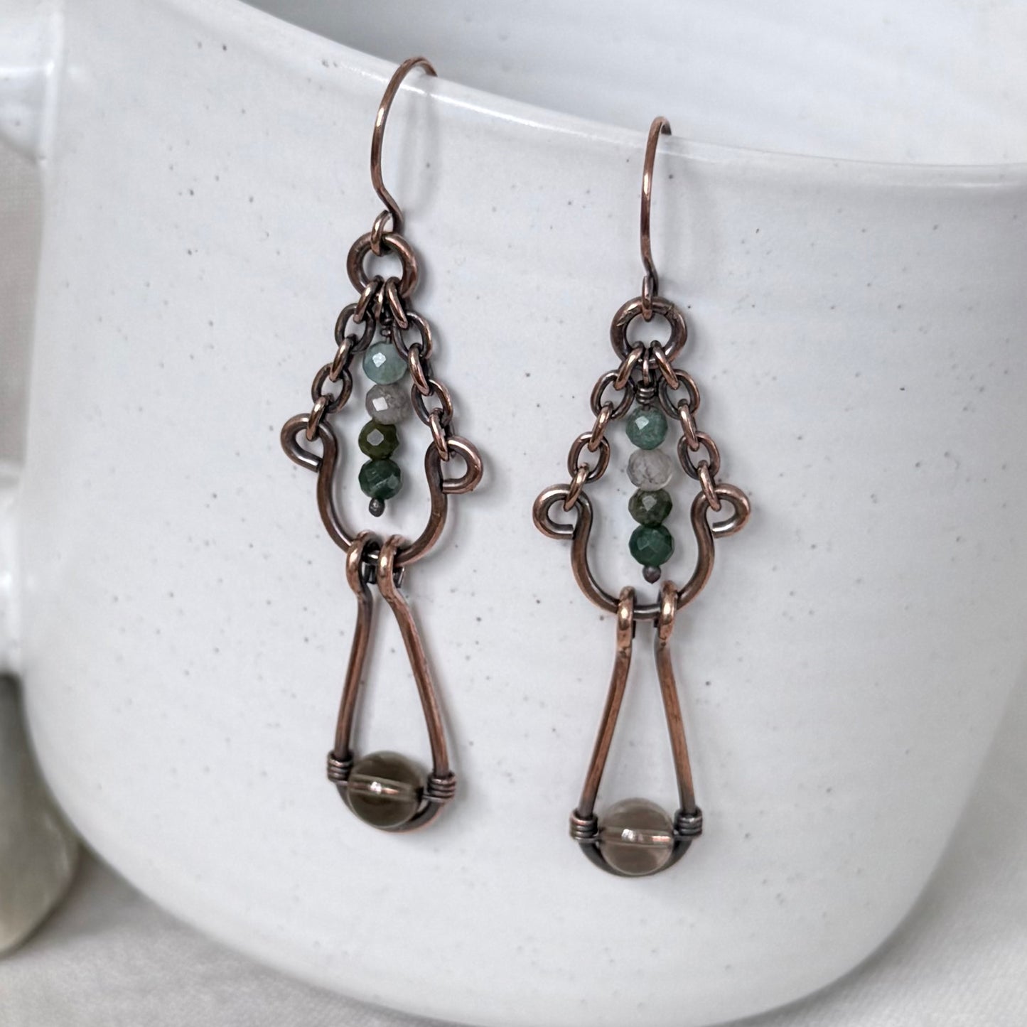 A pair of handmade copper dangle earrings with small green and white beads, and a larger brown bead at the bottom, displayed against a white speckled ceramic mug.