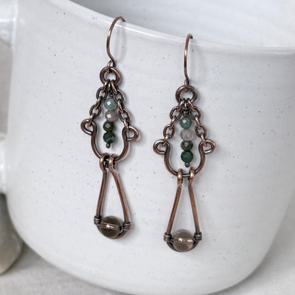A pair of handmade copper dangle earrings with small green and white beads, and a larger brown bead at the bottom, displayed against a white speckled ceramic mug.