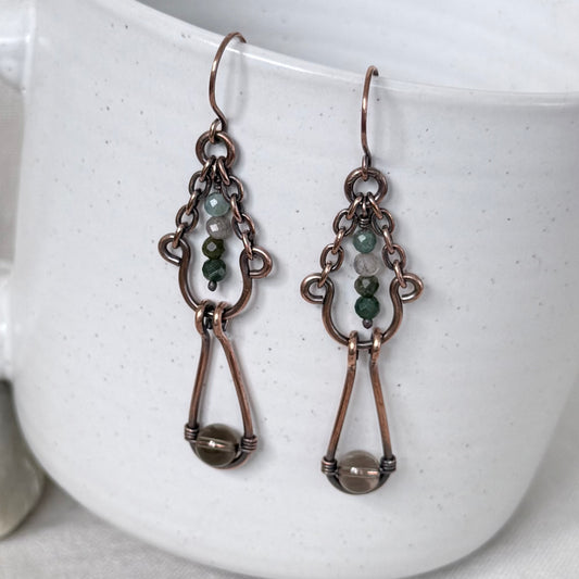 A pair of handmade copper dangle earrings with small green and white beads, and a larger brown bead at the bottom, displayed against a white speckled ceramic mug.
