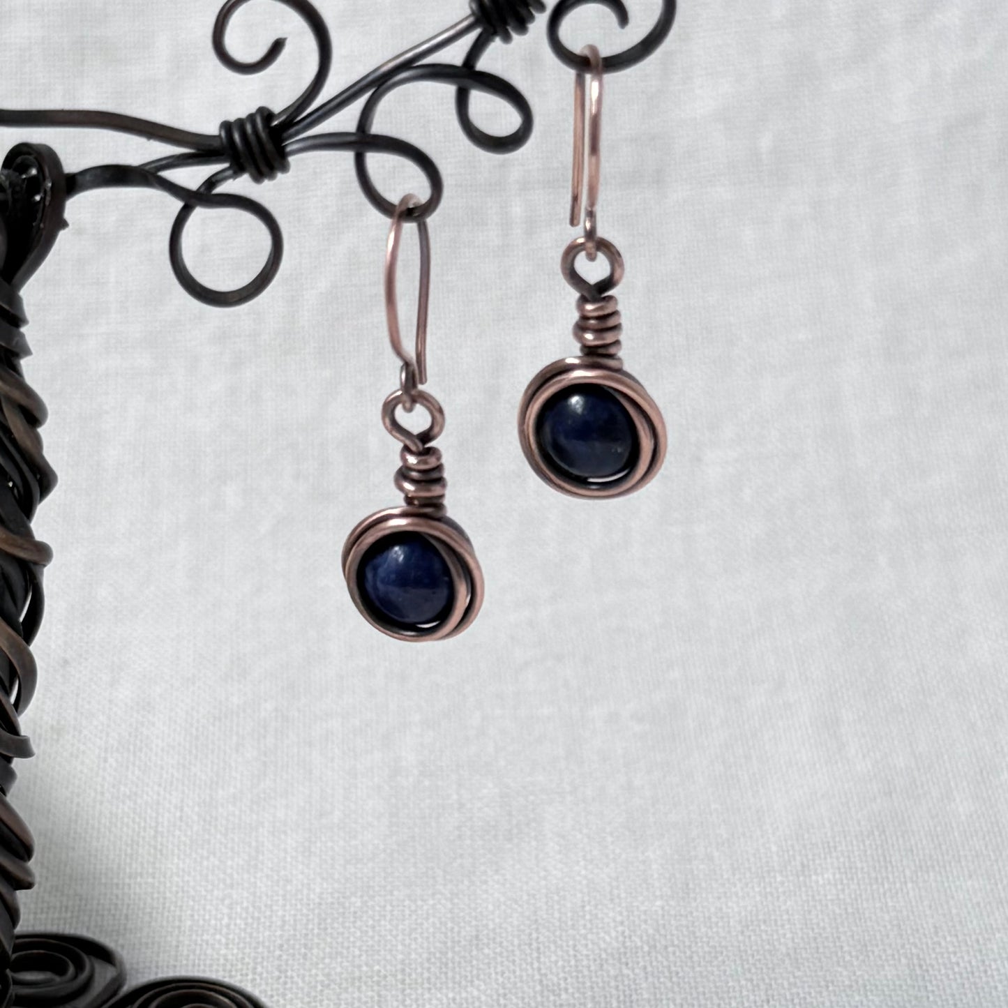 A pair of handmade copper wire-wrapped drop earrings with dark blue round gemstones, hanging from a black metal display stand against a white textured fabric background.