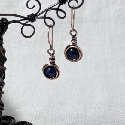 A pair of handmade copper wire-wrapped drop earrings with dark blue round gemstones, hanging from a black metal display stand against a white textured fabric background.