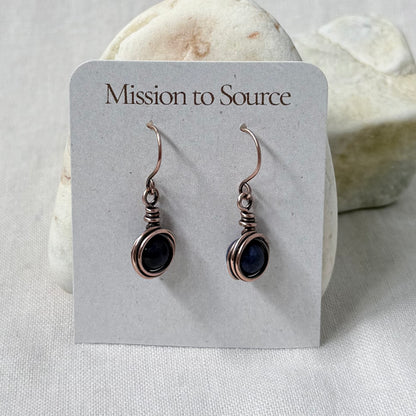 A pair of handmade copper wire-wrapped drop earrings with dark blue stones, displayed on a white card that reads 'Mission to Source' against a soft neutral background.