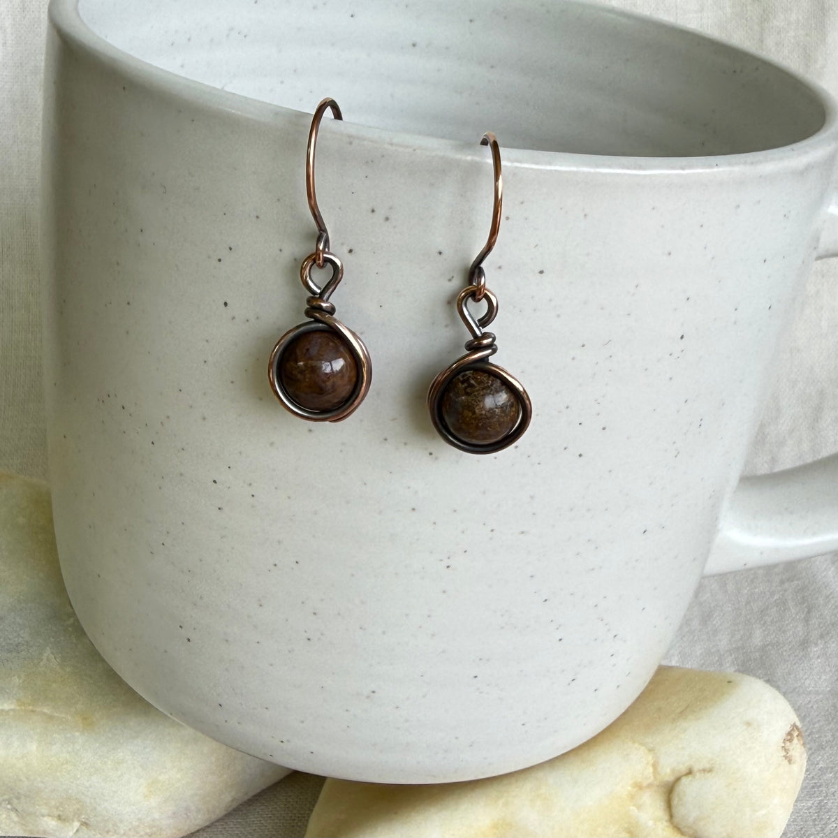 Pair of copper and brown gemstone earrings hanging on a white ceramic cup.